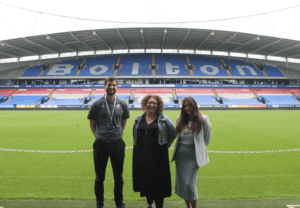 Becci and Amy from Team Boo, standing on the pitch at Bolton Wanderers Football Club with Richard from the BWITC team